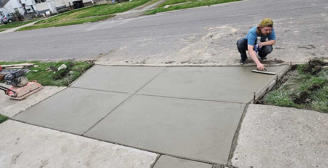 A man in casual attire and a bandana kneels while finishing a freshly laid concrete driveway. The driveway is bordered by wooden forms and connects to an existing sidewalk, with construction tools and a compacting machine on the side.