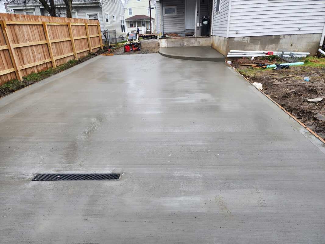 A freshly poured concrete driveway curves gently away from the camera, leading up to a house's side entrance. A strip drain is installed near the foreground, and various construction materials are scattered to the right.