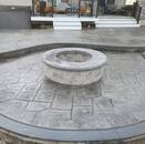 A stamped concrete patio featuring a stone pattern, with a built-in circular fire pit made of paver stones. The patio is adjacent to a house with a porch decorated with pumpkins, highlighting a cozy, outdoor living space.